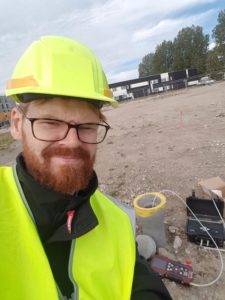 Anton iført hjelm og vest står med fronten vednt mod kameraet. I baggrunden kan man se et prøvetagningsted, hvor et betonrør stikker op ad jorde. Op ad røret er der en slange, hvorfra væske bliver trukket på. Det ligner en byggegrund med sand bagved.