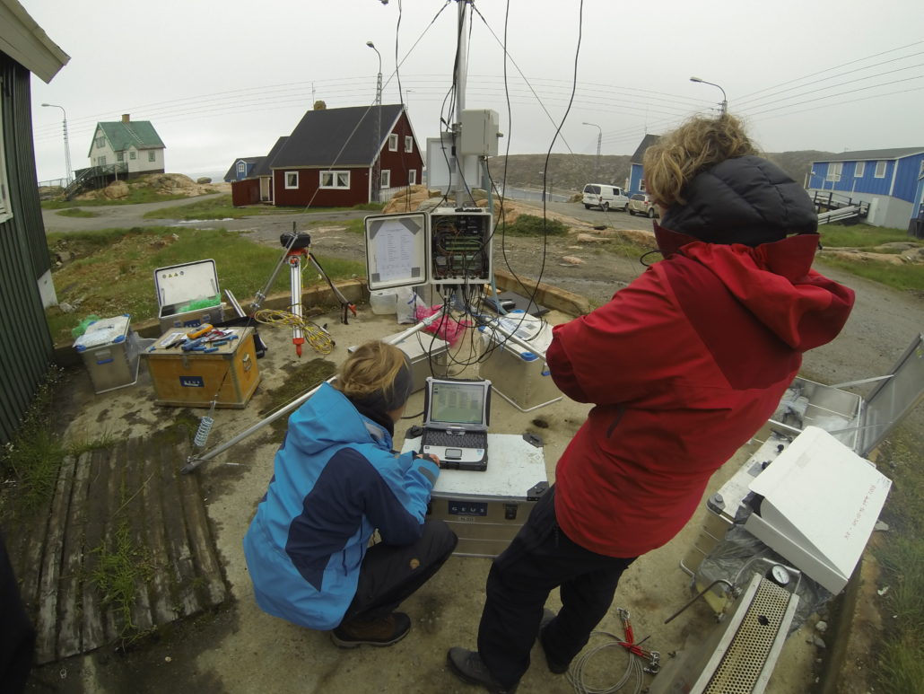 I baggrunden ses et rødt hus i grønland. I forgrundne er to mennesker, den ene er iført blå jakke og sidder på hug ved et apperat. Den anden er iført rød jakke og kigger ned på en maskine.