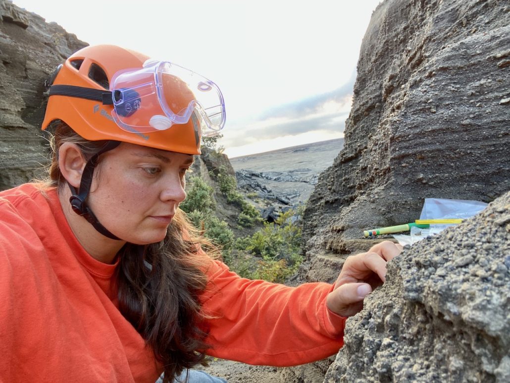 Johanne Schmith iført orange trøje, hjelm og sikkerhedsbriller kigger undersøgende på lag af tefra.