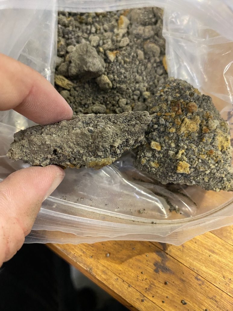 Et stykke af tefra holdes af fingre over en pose med flere stykker indsamlet tefra på et ræbord.