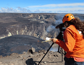 Johanne Schmith str og kigger i en laserafstandsmåler der peger ned på en lavasø i et krater på en skjoldvulkan.