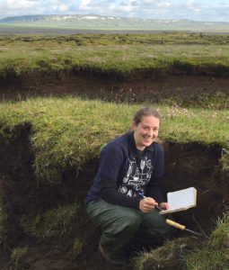 Johanne Schmith sidder i et hul i jorden med en åben blok i den ene hånd og en kuglepen i den anden. Hun sidder på hug og smiler til kameratet. bag hende kan man se grøn jord og bjerge i island.